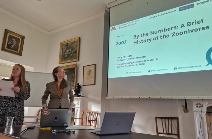 Two researchers standing in front of a projector screen with a title "By the numbers: a brief history of the zooniverse"