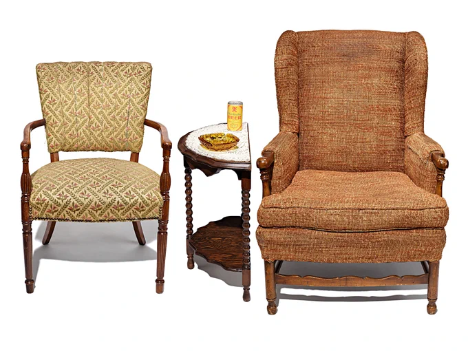 Two fabric-covered living room chairs (the left one a light blonde color, the one on right a medium brown color) with a wooden crescent-shaped side table in between. On the table is a can of beer and an ashtray.