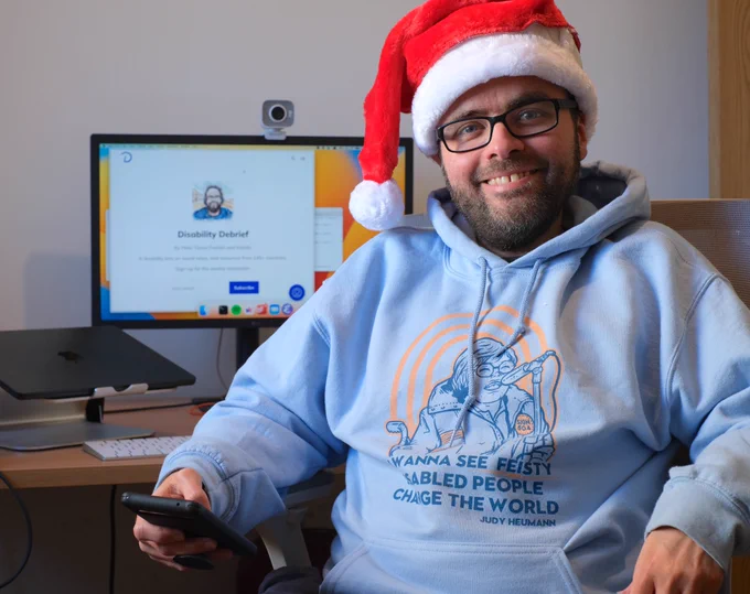 Picture of Peter at his desk, smiling and wearing a Santa hat. Peter is white, has glasses and a beard with speckles of grey. He's wearing a Judy Heumann hoodie that says "I wanna see feisty disabled people change the world".