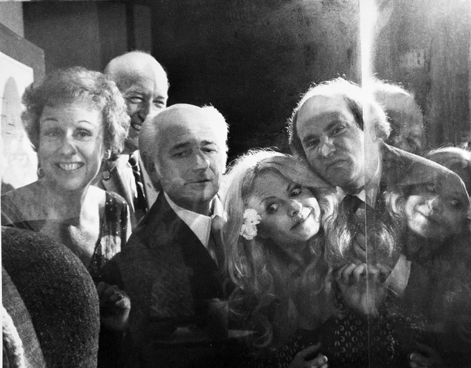 Black and white photo of five adults next to a museum display case, clustered closely together and smiling. They are Jean Stapleton, Smithsonian Secretary S. Dillon Ripley, Norman Lear, Sally Struthers and Rob Reiner as they peer into the case where the chairs are displayed.