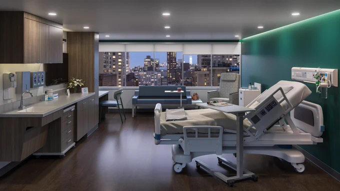 A modern hospital room overlooking a city skyline features warm neutral toned hospital furniture and a bold green accent wall - The Steelcase Empath medical recliner chair, Surround Sleeper Sofa, Convey Modular Storage system, Convey Cart and Radia chair complete the space.