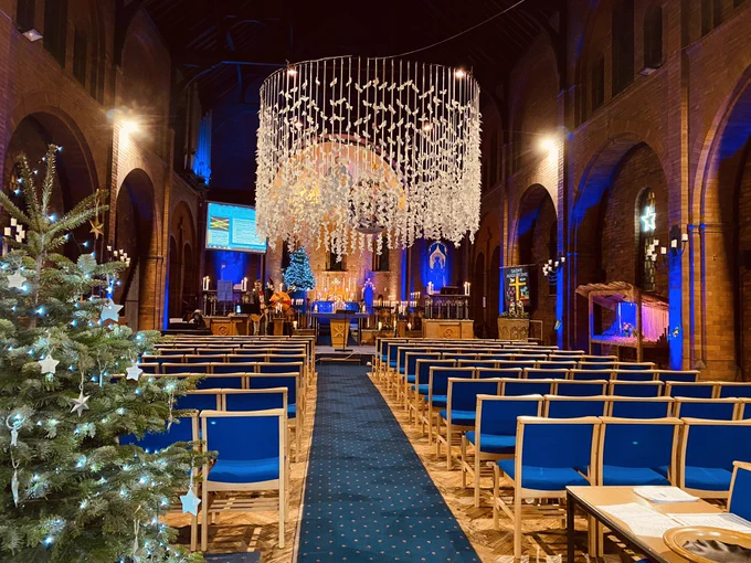 Redbrick church interior with rows of chairs. The church is lit in different colours with lots of candles around. There is a Christmas tree with stars hanging on it. On one side there is a wooden crib with figures of Mary and Joseph placed on straw bedding. Hanging from the centre of the church are over 2000 paper doves on ribbons as a circular display.