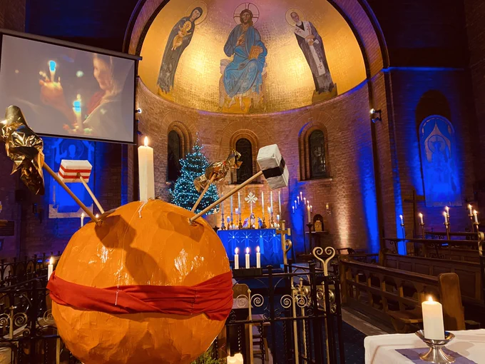A large papier mache christingle is on the left. There are lots of lit candles in the background. A large dome shaped apse is visible with religious artwork painted inside.