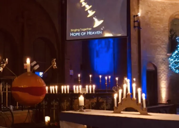 An altar has a candle arch with 7 lit candles. There is a large spherical orange Christingle on the left. In the background is a projector screen with the words “Singjng together. Hope of heaven” written on it.