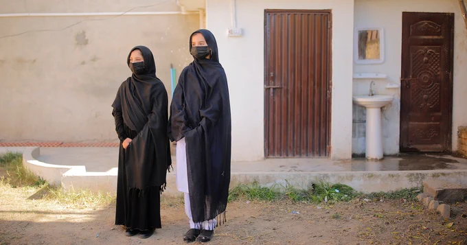 Two young Pakistani girls wearing long black scarfs that are draped over their heads and shoulders stand side-by-side in front of a toilet block. A basin with a shelf and mirror above it can be seen between two closed wooden doors that make up the toilet facilities behind them. Photo credit: IRC Pakistan.