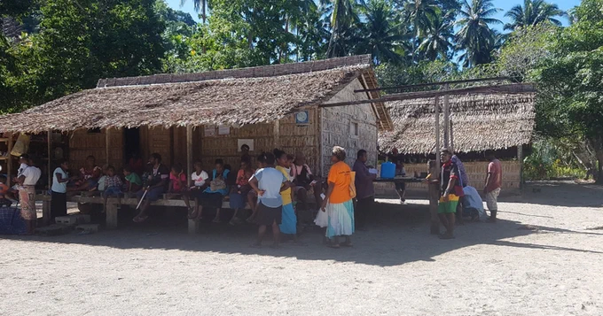 Community members gather in a rural village in western Guadalcanal, Solomon Islands. In the surrounds there are houses made from bamboo with thatched roofs and sandy ground. Photo credit: Plan International Solomon Island and Live & Learn Environmental Education Solomon Islands. 