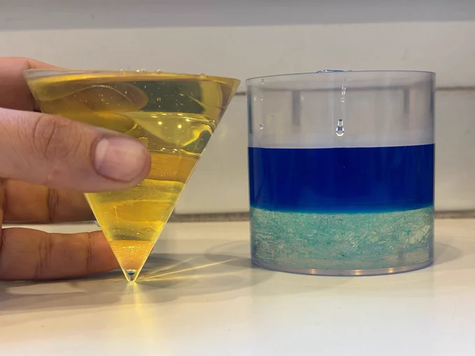 The third photo in the sequence. The cylinder is two-thirds full, with a layer of the clear syrup and a layer of the blue liquid. The cone is filled to the top with yellow cooking oil.