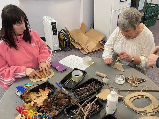 Two ladys sat at a table, one with black hair and a pink jacket, and another with grey hair and a white top. They’re crafting at a table. There are woodland objects (sticks, leaves, cardboard pumpkins).