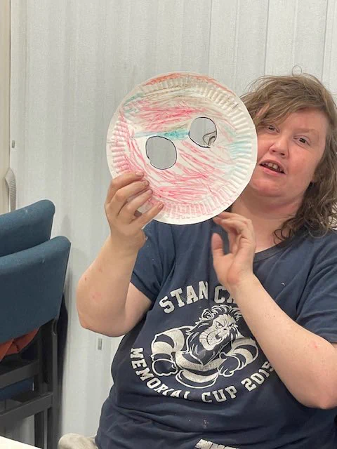 A lady wearing a blue t-shirt, and holding up and mask with two holes in. It’s coloured in red and blue.