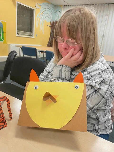 A lady (blonde hair, glasses), sat behind a card she has made. It has a yellow bird on it, with goggly eyes.
