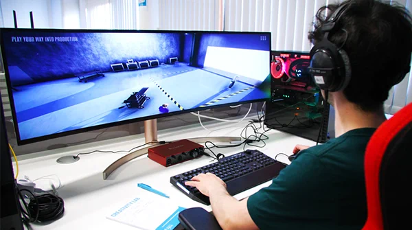 A person wearing headphones sits at a desk with a large curved computer screen showing footage of the play your way into production game. They reach out a touch a keyboard on the desk.