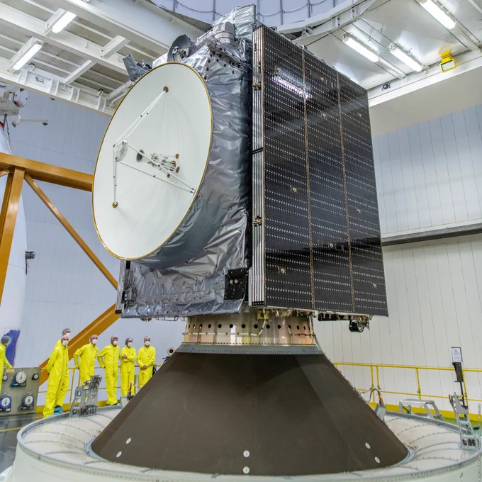 Le satellite Juice repliée et en cours d'intégration sur Ariane 5. C'est un cube, ses panneaux solaires sont repliés sur 2 côtés opposés et une grande antenne est visible sur une troisième face. Une demi douzaine de personnes en combinaison de protection jaune, charlottes et masques, sont visibles à l'arrière plan. Ils semblent minuscules sans qu'on puisse vraiment définir si c'est à cause de la distance ou de la taille du satellite.