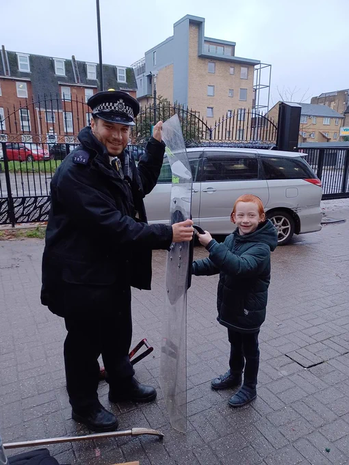 A police officer in uniform and a young Jewish boy who is wearing a kippah are smiling whilst both holding up the same police riot shield.