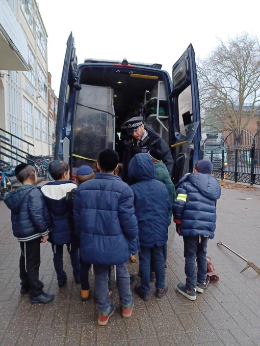 A police officer in uniform is standing in front of a navy blue police van and engaging with eight young Jewish children who are wearing kippahs and are gathered around the officer.