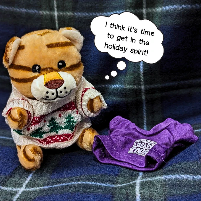 A tan plush cat with brown stripes sits on a blue and green plaid blanket, next to a purple Nassau Library Tour t-shirt. He's wearing a white and red holiday sweater, and has a thought bubble above him that reads "I think it's time to get in the holiday spirit!"