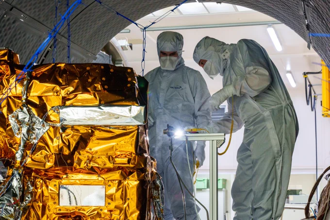 MicroCarb, entouré de couverture thermique doré, est placé dans un grand cylindre en salle blanche. 2 personnes en combinaison intégrale, masque et gants sont en train de l'examiner : le satellite est un peu plus petit qu'eux.