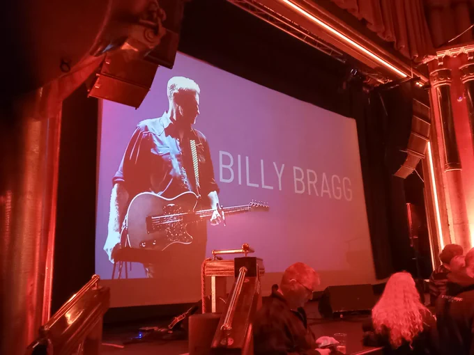 A red-lit stage area with a backdrop screen saying "Billy Bragg " and a muted colour image of him with a guitar around his neck.