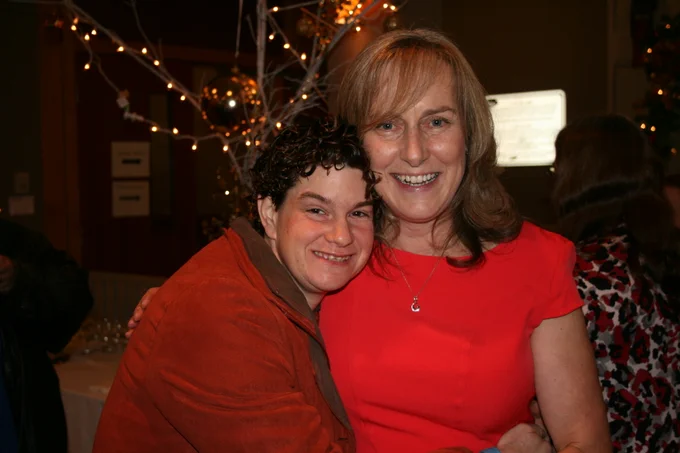 Two women stand side by side with their arms around each other. They are both wearing red and smiling at the camera.