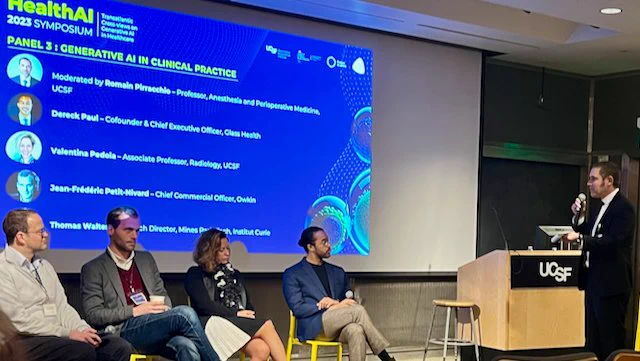Four persons are sitting as part of a panel. One moderator is standing at a podium. The blue screen says Health AI Symposium 2023 with the names of the speakers. 