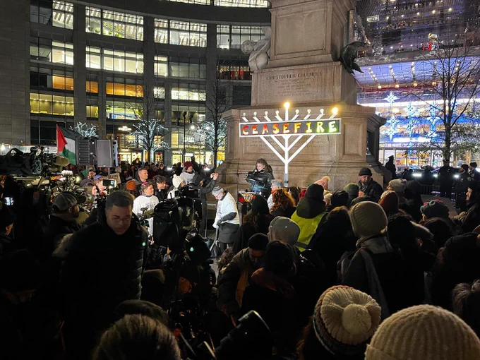 A large crowd of people gathered in front of a statue. In front of the statue is a tall chanukiah with the word CEASEFIRE on it.