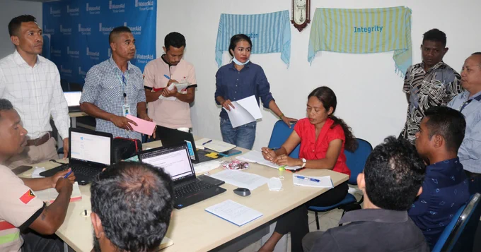 WaterAid, HAK and partner organisation team members seated at and standing around a table with open laptops and documents. They are discussing the review of the Community Score Card indicators. Photo credit: WaterAid / Antoneta Soares.