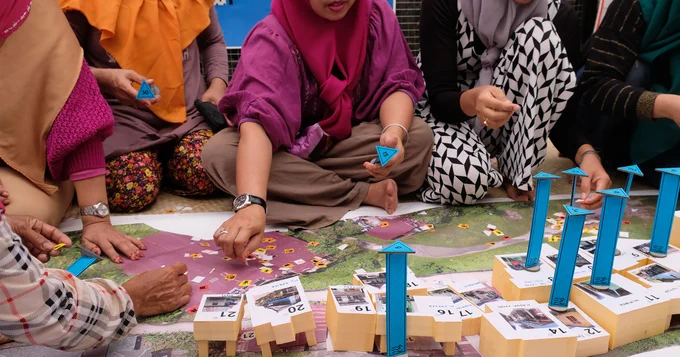 A group of Indonesian women seated cross-legged on the floor attaching pieces of paper to an aerial picture of an urban area and with stacked blocks representing buildings and infratructure in the foreground. They are participating in a PANRITA design workshop in an informal settlement in Makassar, Indonesia. Photo credit: Monash University.