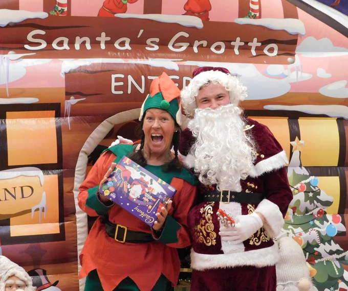 Santa and an elf standing in front of his grotto, holding chocolate selection boxes.