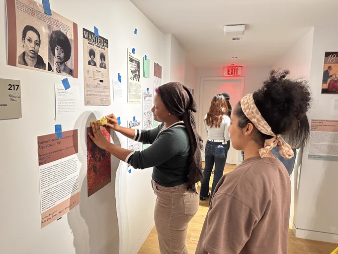 A Black woman wearing a brown scarf wrapped around her head, a green sweater, and khakis hanging up a piece of art on the wall