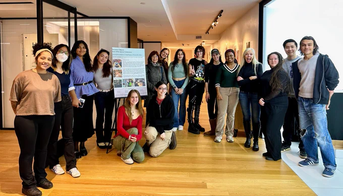  In an art studio-like space, a row of students with two faculty stand and smile with two girls kneeling in front of a poster board on an easel