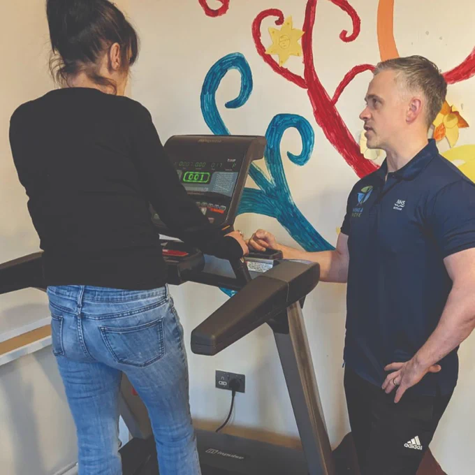 Patient walking on treadmill, supervised by a member of staff. The equipment is in front of a brightly coloured wall.