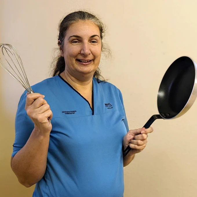 Woman health worker in blue NHS Scotland uniform holding a whisk and a frying pan.