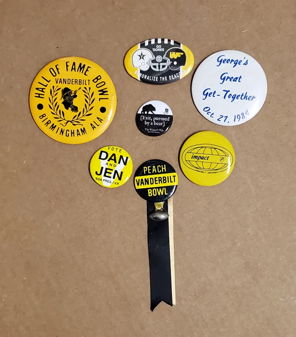 Yellow button with black text, "Hall of Fame Bowl / Vanderbilt / Birmingham Ala." Yellow, white, and black button with text, "Go 'Dores / Demoralize the Deacons". White button with blue text, "George's Great Get-Together / Oct. 27, 1984". White and black button with text, "[Exit, pursued by a bear] / The Winter's Tale / Act III, Scene I". Yellow and white button with black text, "Vote Dan and Jen SGA Pres. / V.P." Yellow button with black insignia of the Impact Symposium at Vanderbilt University. Yellow and black button with a black and gold ribbon and a silver football charm, with text, "Peach / Vanderbilt / Bowl".