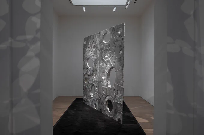 Image: A silver architectural sculpture with holes throughout the square, sheet-like shape, stands in a gallery space. 