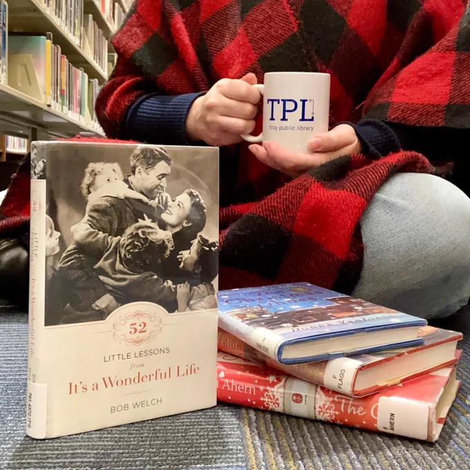 It's a Wonderful Life book cover in front of person wrapped in plaid blanket with white, Troy Public Library coffee mug