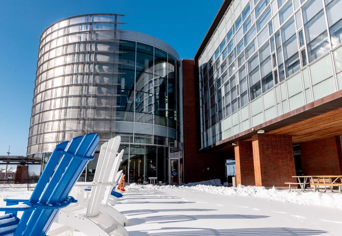 In front of the North Oshawa Campus Library on a clear winter day. The blue, orange and white muskoka chairs that line the reflecting pool are on the left side and you can see a blanket of snow on the ground in front of the library. 