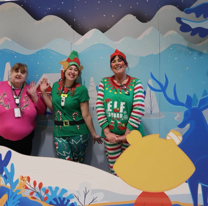 Three females stand smiling in an interactive winter wonderland scene. The scene is shades of blue with a yellow Theo Bear cartoon in the right lower corner.