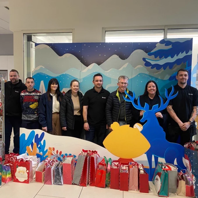 A group of people stand in a winter scene. In front of the scene there are Christmas gift bags.