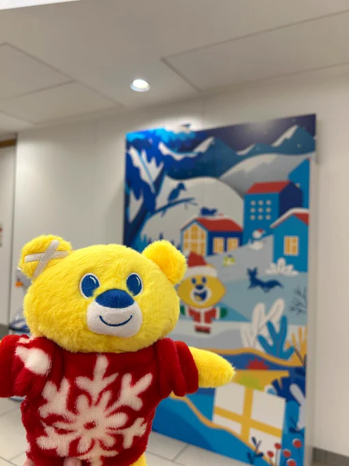 A yellow teddy bear with a red Christmas jumper on stands in front of a blue The Children's Hospital Charity branded cardboard backdrop.