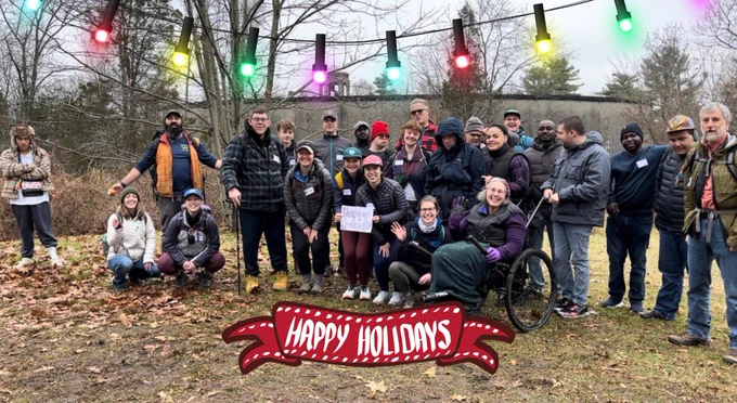 ID:  A group of 20  adults and young adults wearing winter gear standing or  sitting on a grassy ground holding up a sign that says "Happy Holidays".  There is graphic addition of holiday colored lights above and a red  "Happy Holidays" banner.