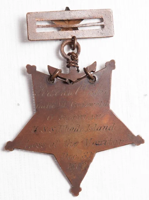 Five pointed bronze star tipped with trefoils containing a crown of laurel and oak. In the center is Minerva, the Roman goddess of wisdom and warfare, standing with left hand resting on fasces and right hand holding a shield blazoned with the escutcheon of the United States. She repulses discord, represented by a male figure holding snakes. It is made of solid red brass, oxidized and buffed.