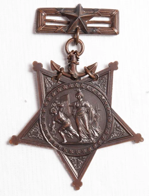 Five pointed bronze star tipped with trefoils containing a crown of laurel and oak. In the center is Minerva, the Roman goddess of wisdom and warfare, standing with left hand resting on fasces and right hand holding a shield blazoned with the escutcheon of the United States. She repulses discord, represented by a male figure holding snakes. It is made of solid red brass, oxidized and buffed.