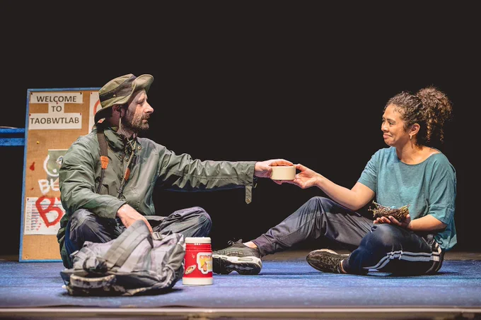 A production shot from The Mother's Return, a theatre piece at the Ludlow Assembly Rooms. A middle-aged, white man with brunette hair and a beard wears outdoor gear; a dark green raincoat, a khaki rain hat and black waterproof trousers. The other is a young, mixed-race woman with curly brown hair in a ponytail wearing running gear; a blue top, black running trousers and trainers. The man is offering a cup of tea out of a red thermos to the woman. She holds a bird’s nest in her other hand.