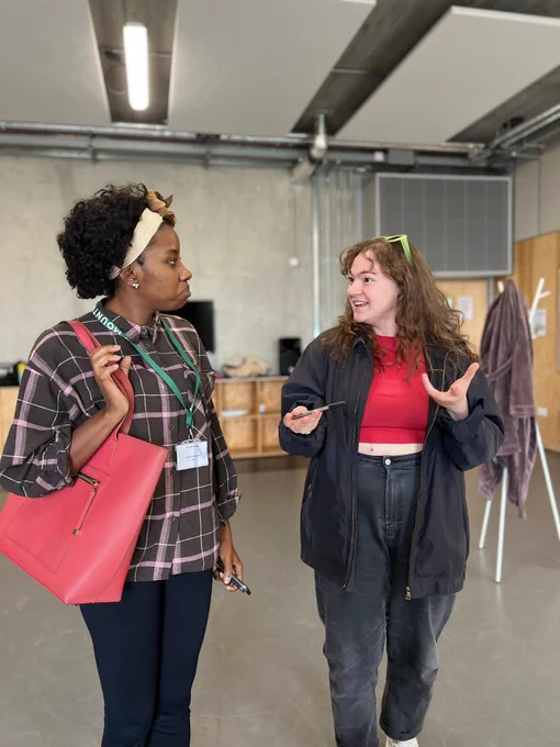 A rehearsal shot from Stick It to Me, a theatre piece at View Festival 2023: A young, black women with curly black hair with a white head band stands wearing a purple checked shirt, black leggings, and a green lanyard around her neck. She has a red bag over her shoulder. She is looking at a young, white brunette girl who is wearing a red top, black jeans, an oversized jacket, and lime green sunglasses on her head.