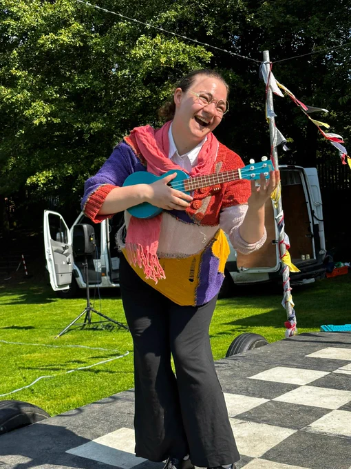 A production shot from Bad Dad, an outdoor touring theatre show: A young, white female with brunette hair in a tight bun stands on a black and white checkered stage. She wears a brightly coloured pink, purple and yellow knitted jumper, black trousers, and round glasses. She holds a blue ukulele.