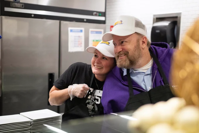Two people pose smiling behind a food counter.