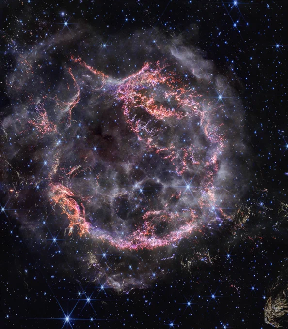 A roughly circular cloud of gas and dust with complex structure. The inner shell is made of bright pink and orange filaments studded with clumps and knots that look like tiny pieces of shattered glass. Around the exterior of the inner shell, there are curtains of wispy gas that look like campfire smoke. Around and within the nebula, various stars are seen as points of blue and white light. Outside the nebula, there are also clumps of dust, coloured yellow in the image.