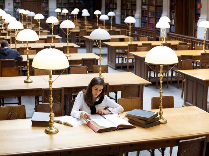 Person sat at a desk reading a book, with other books surrounding them.