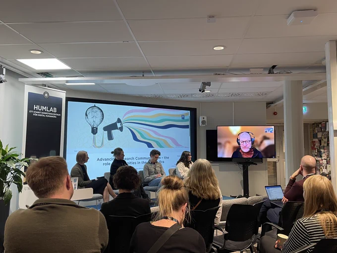 Members of a panel are ready to start the panel discussion. The members are sitting on a small stage, and one member is online, on the screen. The audience is looking at them. 