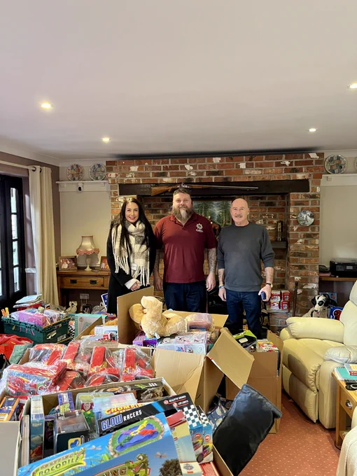 two members of staff and a volunteer for Tara's Angels stand inside a living room, surrounded by boxes of children's toys