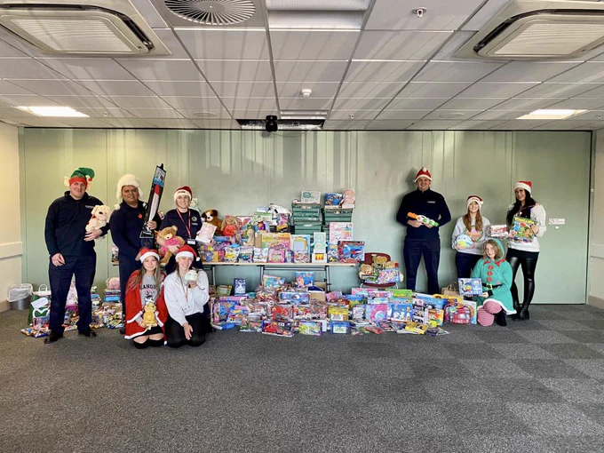 a group of staff stand/sit next to a huge pile of children's toys, wearing santa hats